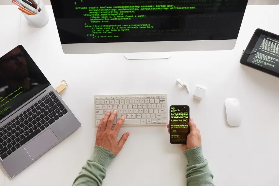Developer la birou cu cod pe ecrane și smartphone, tastură wireless și mouse alb pe masă.