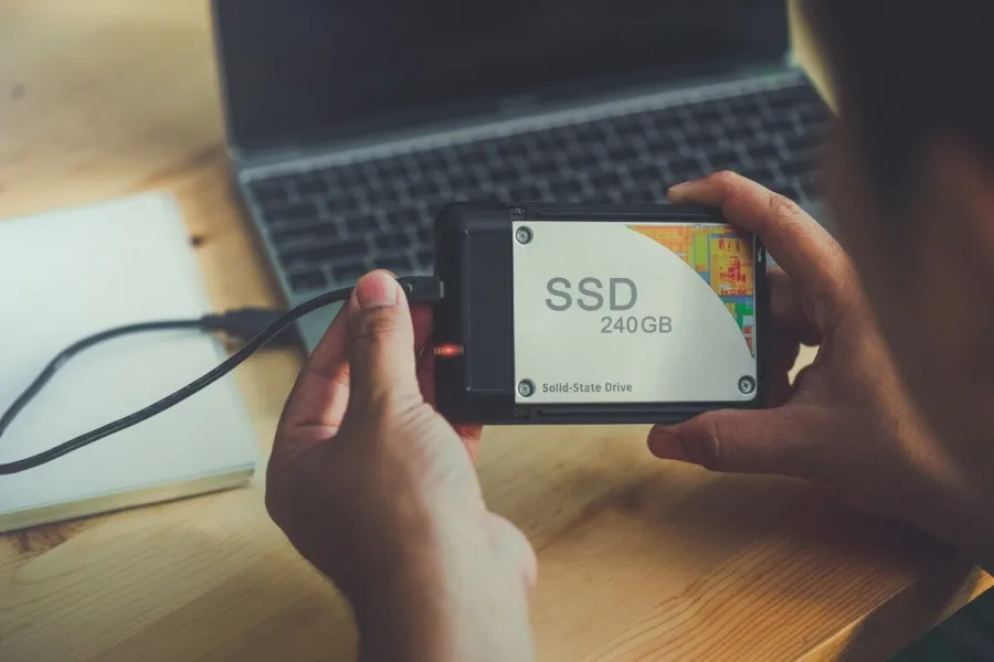 Persoană ținând o unitate SSD 240GB cu cablul conectat, laptop în fundal pe birou din lemn